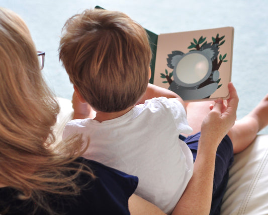 An adult reading a board book with a toddler. The book has a picture of a koala and a mirror in place of the face. 